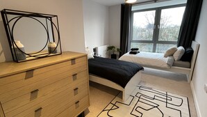 Een moderne slaapkamer met een houten dressoir, een bed met zwart beddengoed, een nachtkastje met een lamp en een groot raam met gordijnen.