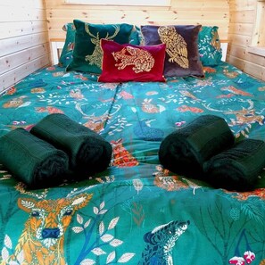 Una cama con una colcha floral de colores, dos toallas enrolladas y almohadones decorativos con motivos de animales.
