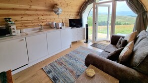 A cozy living room with a sofa, a coffee table, and a TV. There's a kitchen area with a microwave, a kettle, and a bottle. The room has large windows with a view of the countryside.