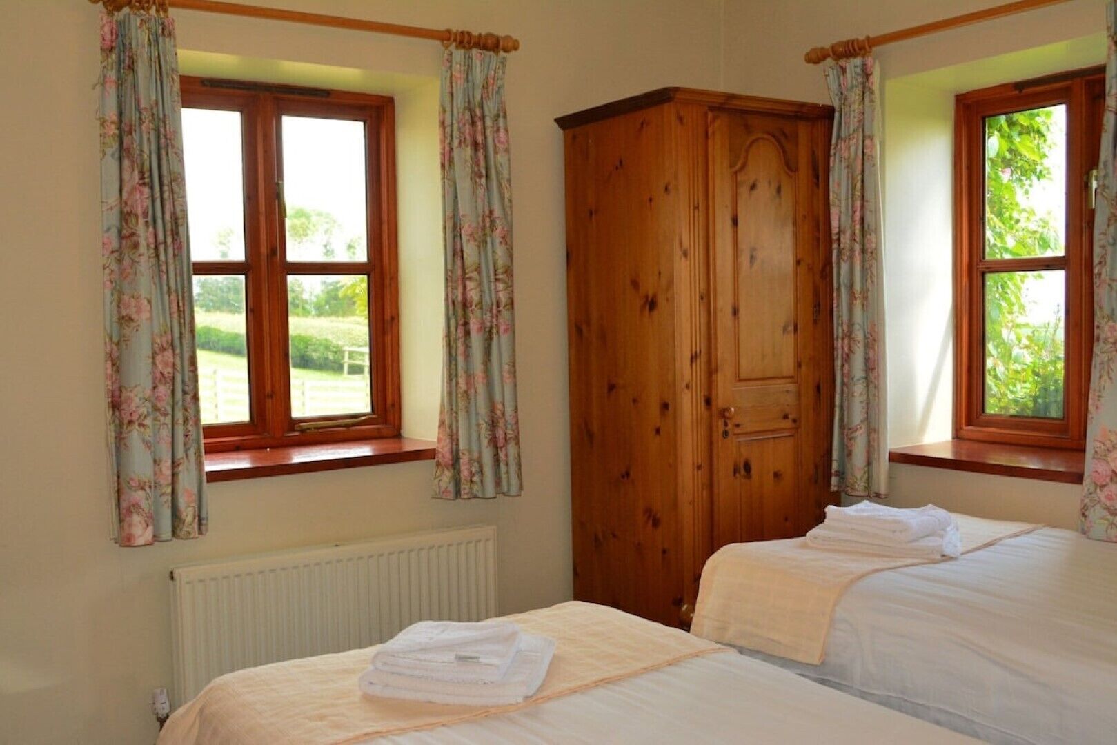 A room with two beds, a wooden wardrobe, and windows with floral curtains.