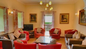 Ein Wohnzimmer mit roten Sofas, einem Glastisch und einem Fernseher. An den Wänden hängen zwei Gemälde, und eine Chandelüster ist angebracht.