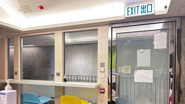 A room with a glass door marked "EXIT DOOR," a blue chair, a yellow chair, a white table with a bottle, and a small black object on the table. - POP E INN (Kowloon)
