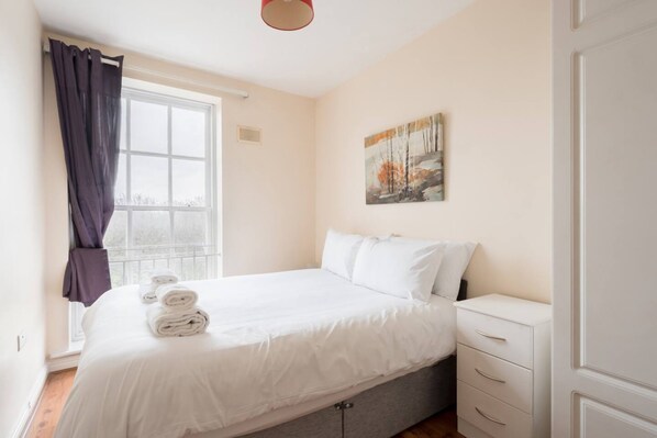 A bedroom with a bed, a window with curtains, a painting on the wall, and a white dresser.