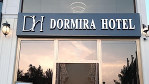 La entrada del Hotel Dormira, con un amplio ventanal de vidrio, una alfombra morada y un letrero en idioma extranjero.