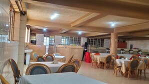 A restaurant with multiple tables and chairs, a kitchen area, and a person standing near the kitchen. - Maralink Hotel (Narok)