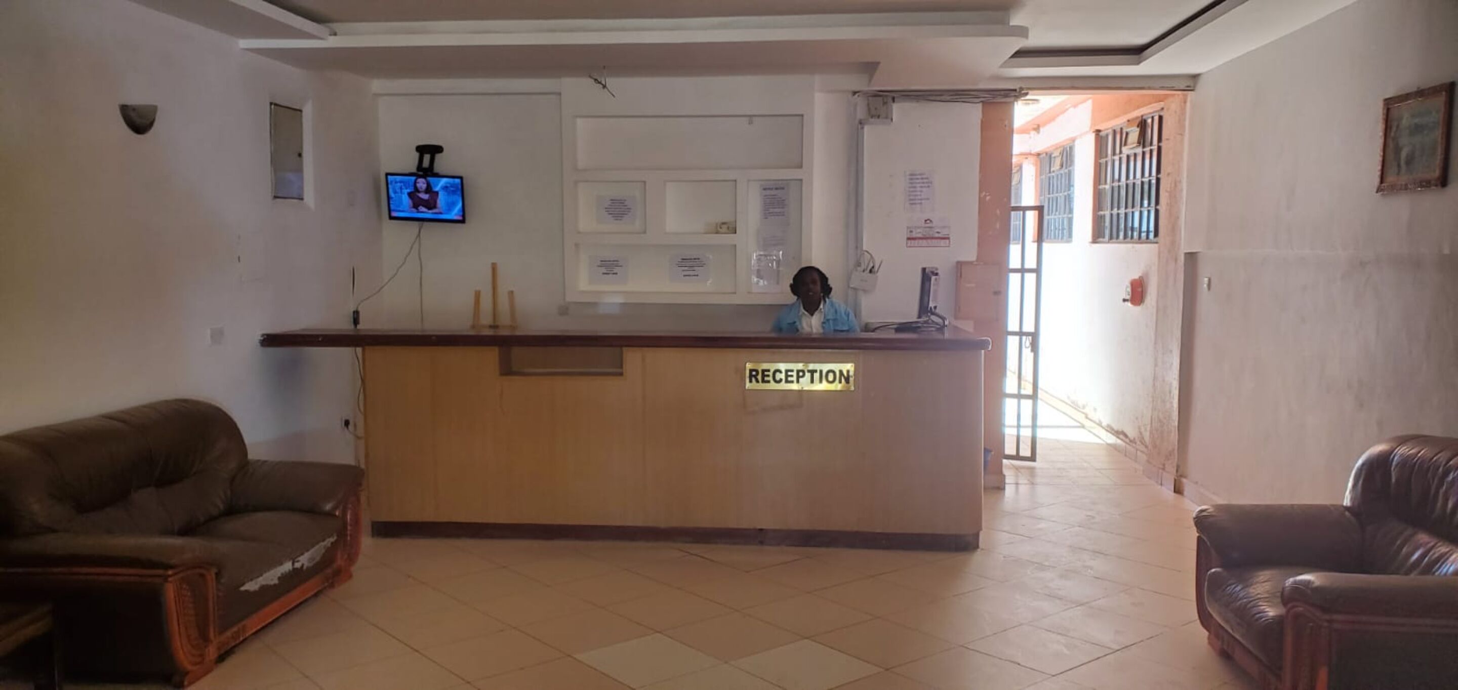 A reception area with a wooden desk labeled "RECEPTION," a television mounted on the wall, and a person standing behind the desk.