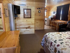 Interior acogedor de una cabaña con pared de madera, una cama con manta de estampado de hojas, una mesa de comedor y una zona de cocina.