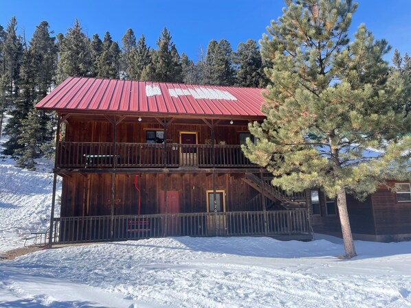 Una cabaña de madera de dos pisos con techo rojo, rodeada de nieve y pinos.