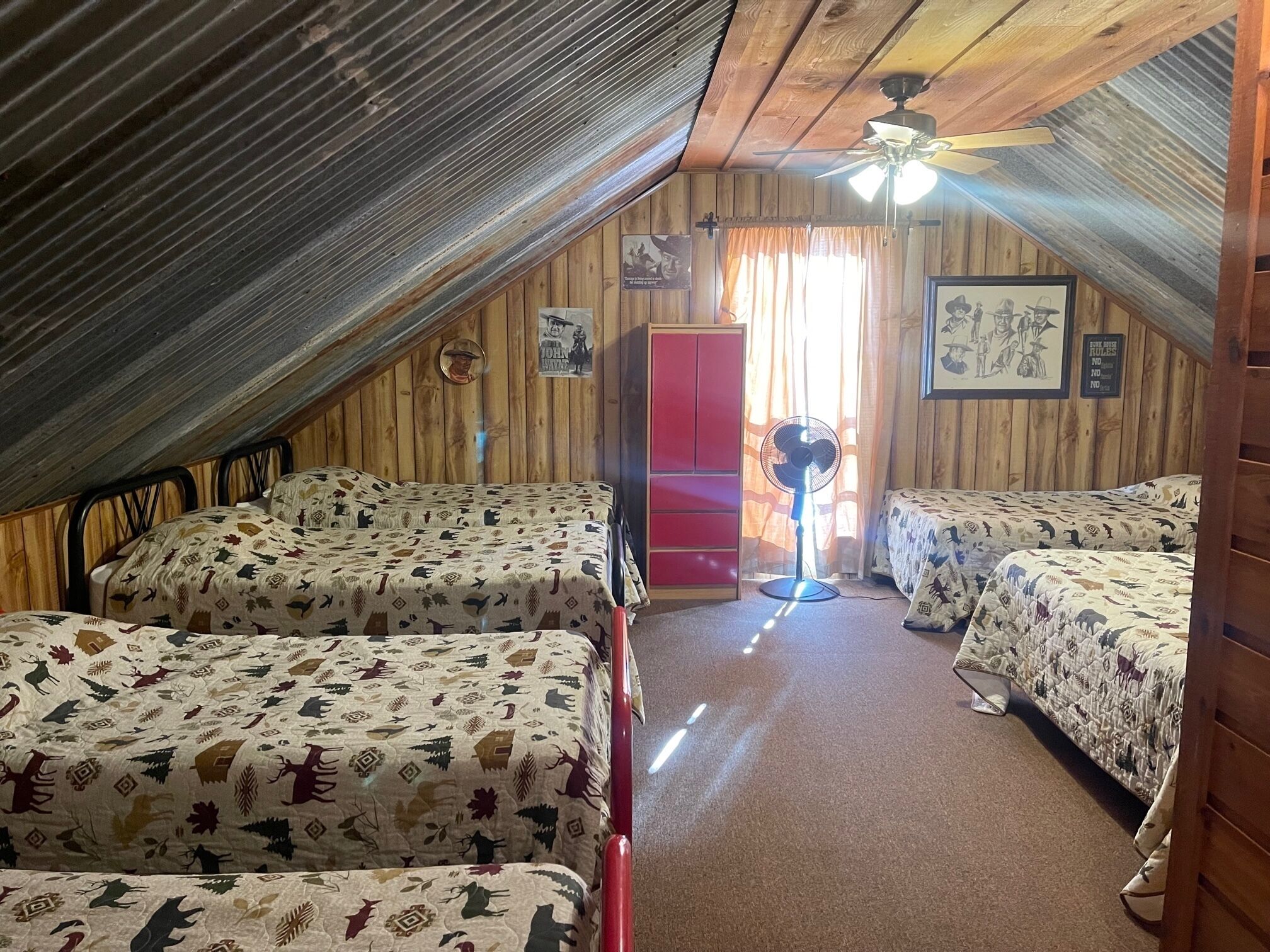 A dormitory room with two beds, a red wardrobe, a ceiling fan, and a window with curtains.