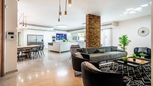 Ein modernes Wohnzimmer mit einem grauen Sofa, schwarzen Ohrensesseln und einem Couchtisch. Im Hintergrund befindet sich eine Küche mit einer weißen Kochinsel und ein Essbereich mit Tisch und Stühlen.