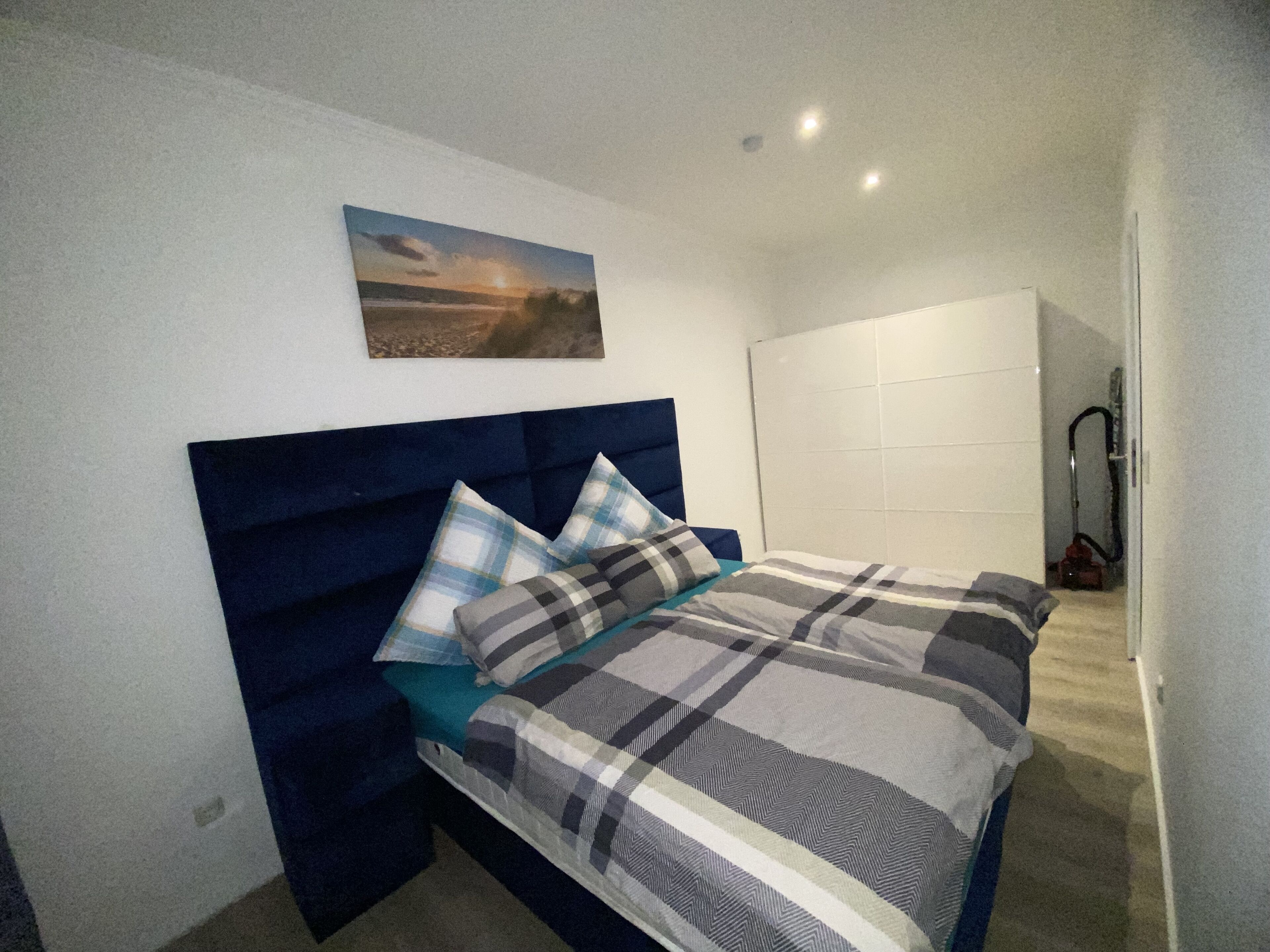 Een slaapkamer met een bed, een hoofdbord, een schilderij en een stofzuiger op de achtergrond.