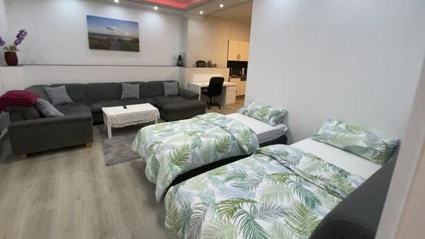 A modern living room with a sofa, two single beds with tropical prints, a coffee table, and a kitchen in the background. - Schöne Wohnung 72qm, Nähe Stadt- & Schulzentrum, 8 km von der Tschechei Entfernt (Selb)