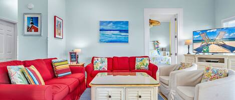 Een woonkamer met een rode bank, een witte salontafel en een beige fauteuil. Aan de muur hangt een schilderij van de zee en er staat een televisie op een dressoir.