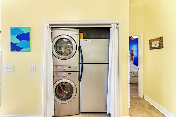 A laundry room with a front-loading washer and dryer, a refrigerator, and a door leading to a hallway with a blue wall and a painting. - Beachfront, Private home, dog friendly, heated pool, 4 BR (St. George Island)