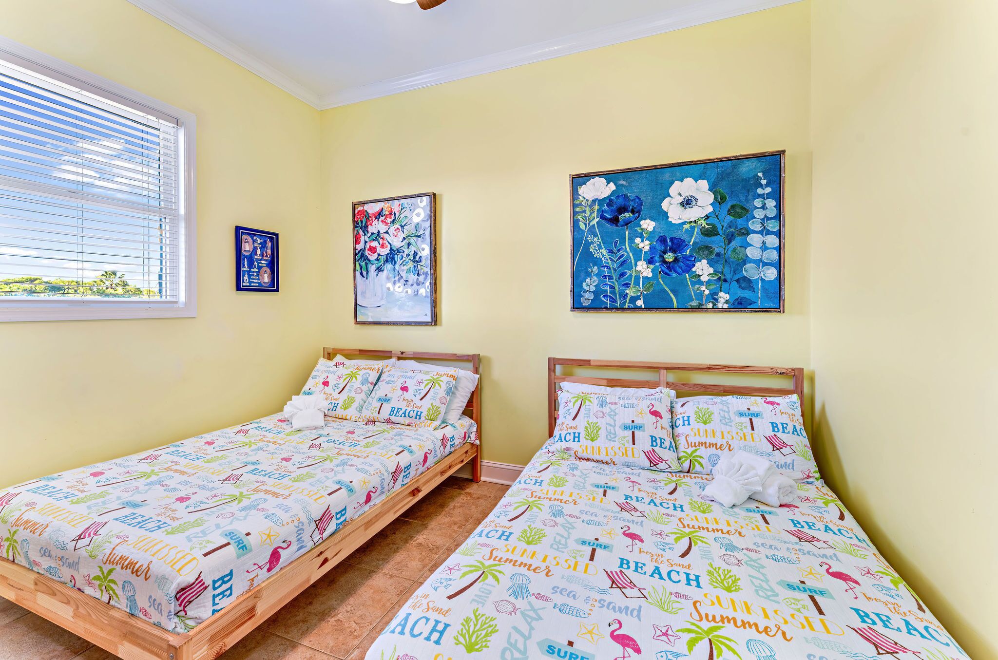 A room with two wooden beds, each with colorful beach-themed bedding. There are two framed floral paintings on the walls and a window with blinds.