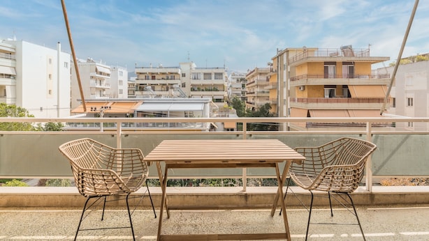 Un balcone con un tavolo di legno e due sedie in vimini, con vista su una zona residenziale con edifici a più piani.