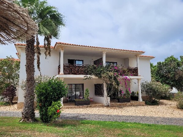 A two-story building with a balcony, surrounded by greenery and a gravel driveway.