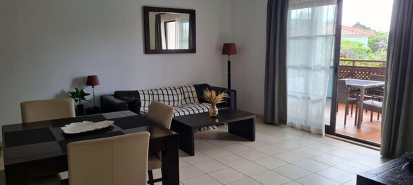 A living room with a sofa, coffee table, and dining table. There is a balcony with a view of greenery.