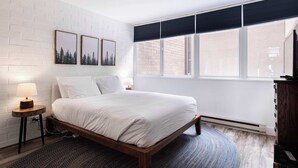 Ein Schlafzimmer mit einem Bett, einem Nachttisch, einer Lampe, einem Fenster mit Jalousien und einer Wand mit drei gerahmten Bildern.