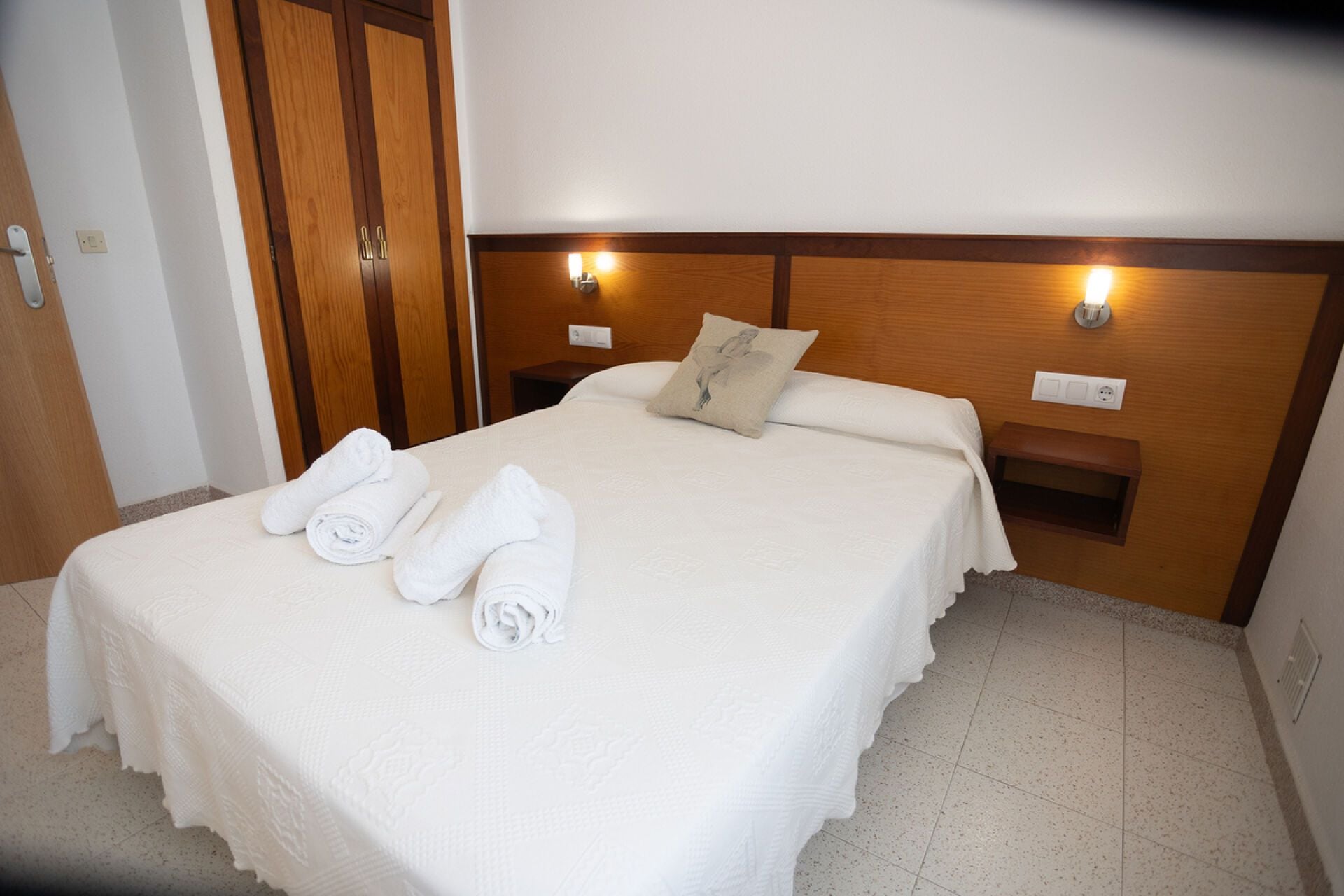 Een hotelkamer met een net opgemaakt bed, twee nachtlampjes en een houten hoofdeinde.