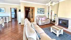 A living room with a beige sofa, a fireplace, a bookshelf, and a dining area with a table and chairs.