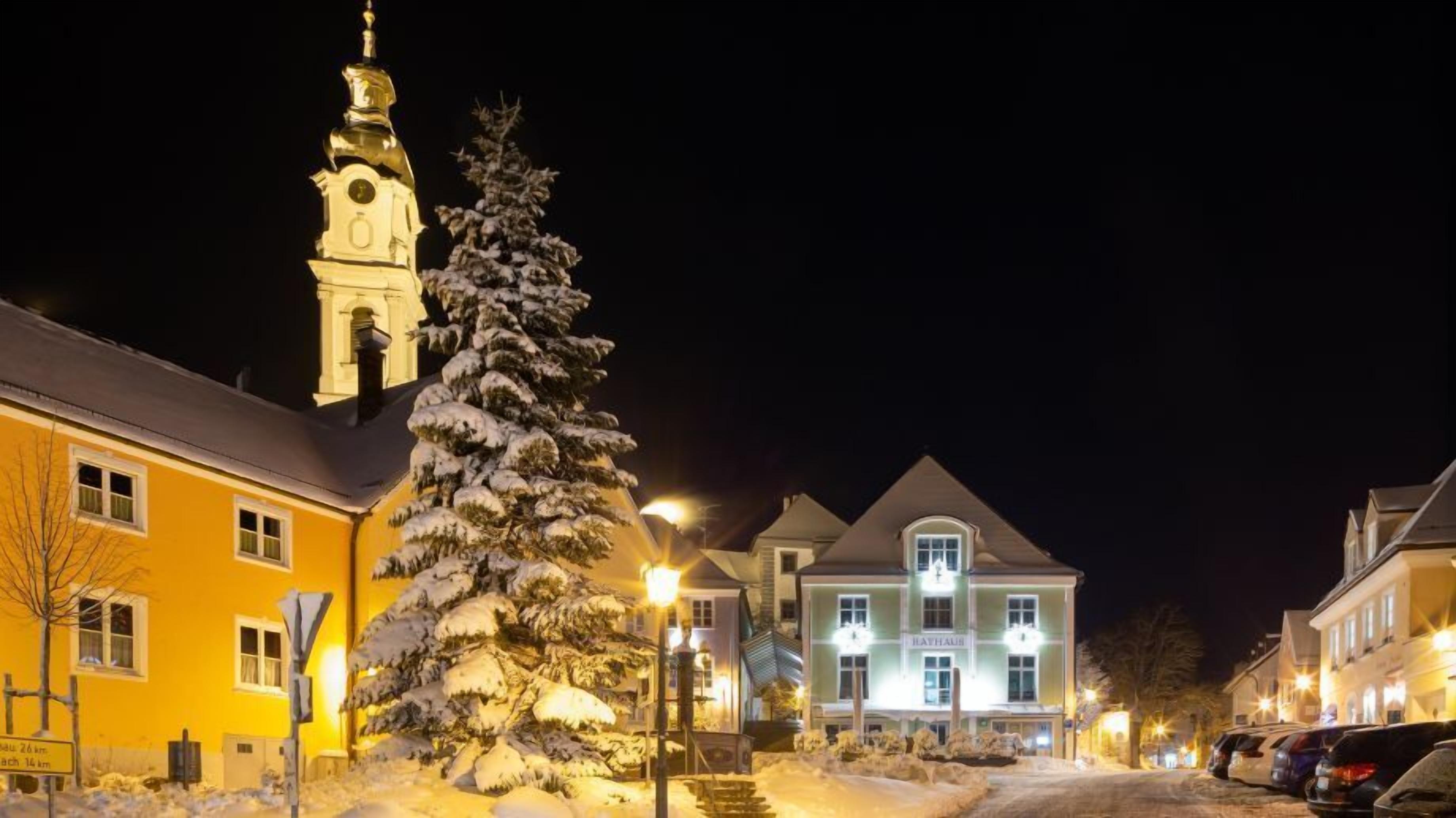 Une rue enneigée, de nuit, avec un clocher d’église, un sapin de Noël et des bâtiments illuminés.