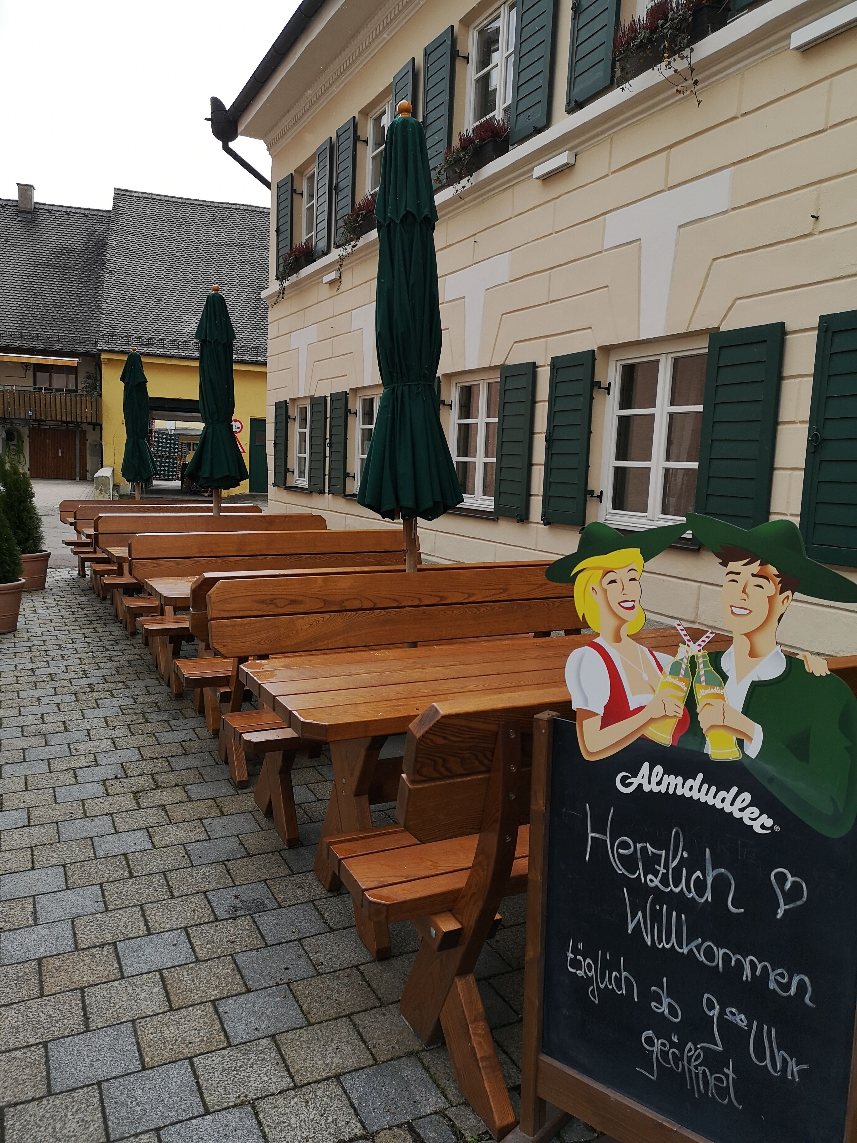 An outdoor seating area with wooden benches and green umbrellas, a chalkboard sign with greetings, and a building with green shutters.