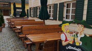 An outdoor seating area with wooden benches and green umbrellas, a chalkboard sign with greetings, and a building with green shutters.