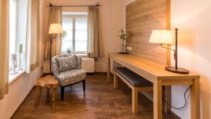 A cozy room with a wooden floor, a patterned armchair, a wooden bench, a table with a lamp, and a window with curtains. - Brauereigasthof & Hotel Maierbräu (Altomunster)