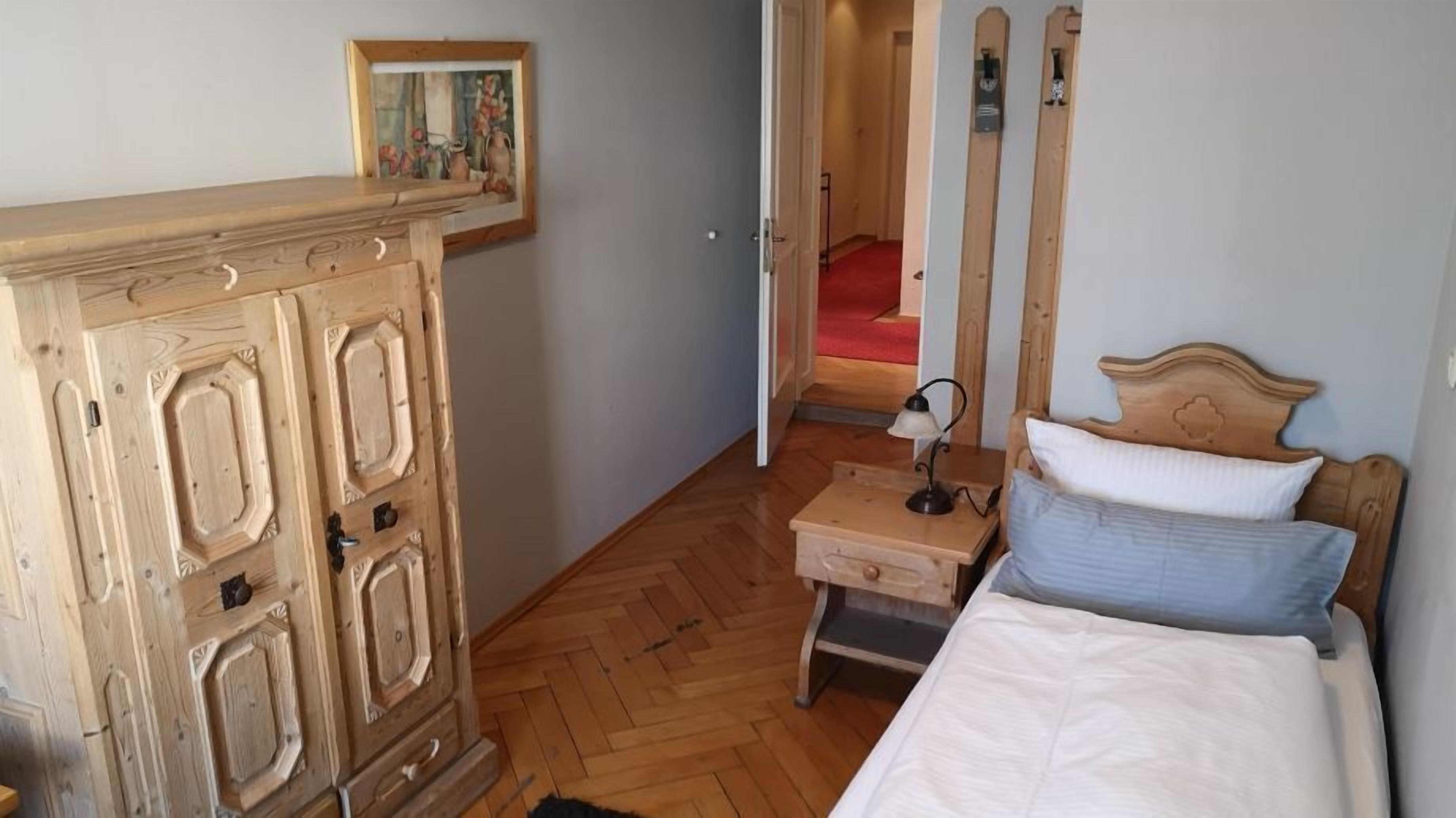 A bedroom with a wooden wardrobe, a bed with pillows, a nightstand with a lamp, and a painting on the wall.