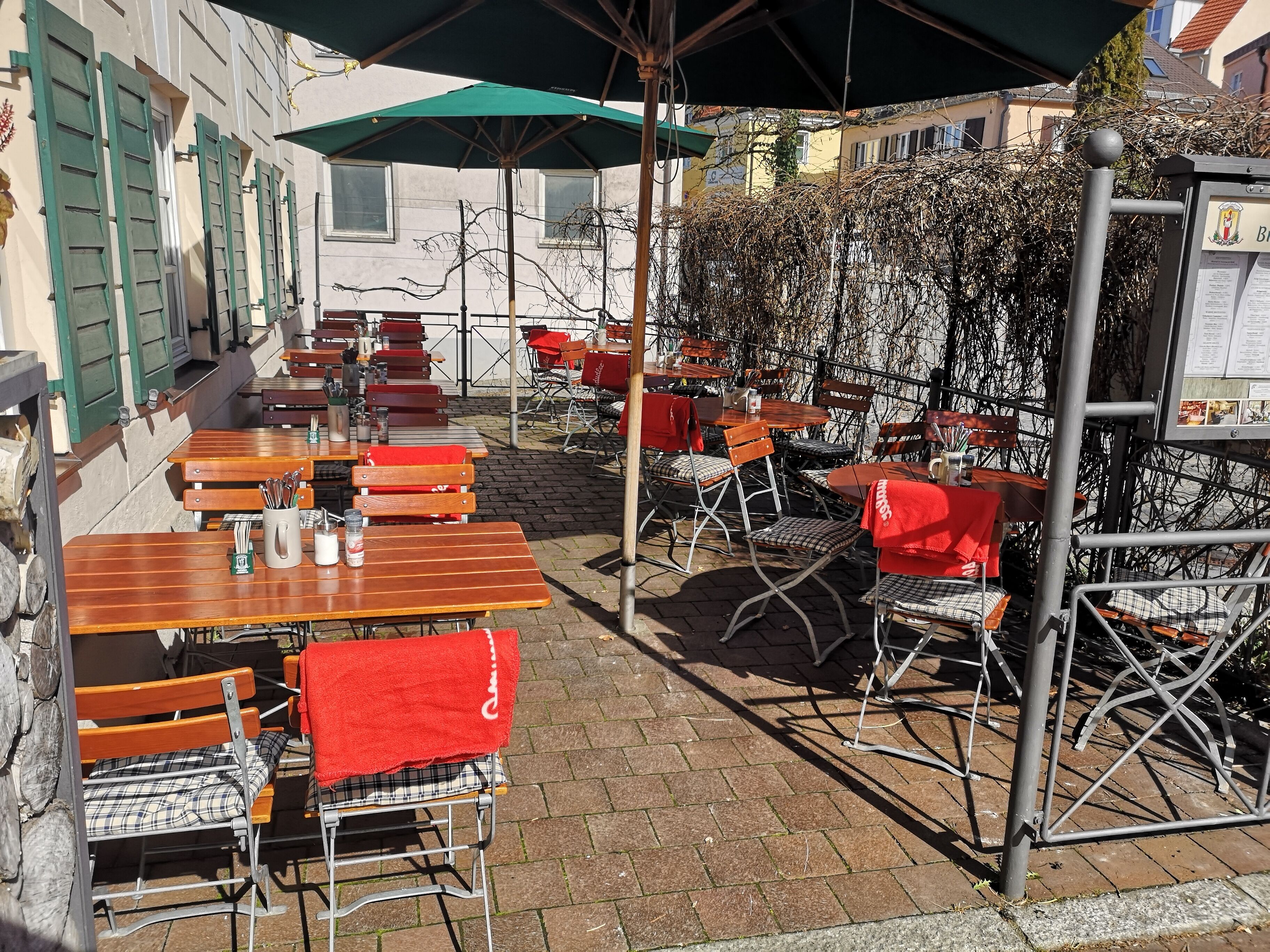 An outdoor café with wooden tables and red chairs, some with umbrellas.