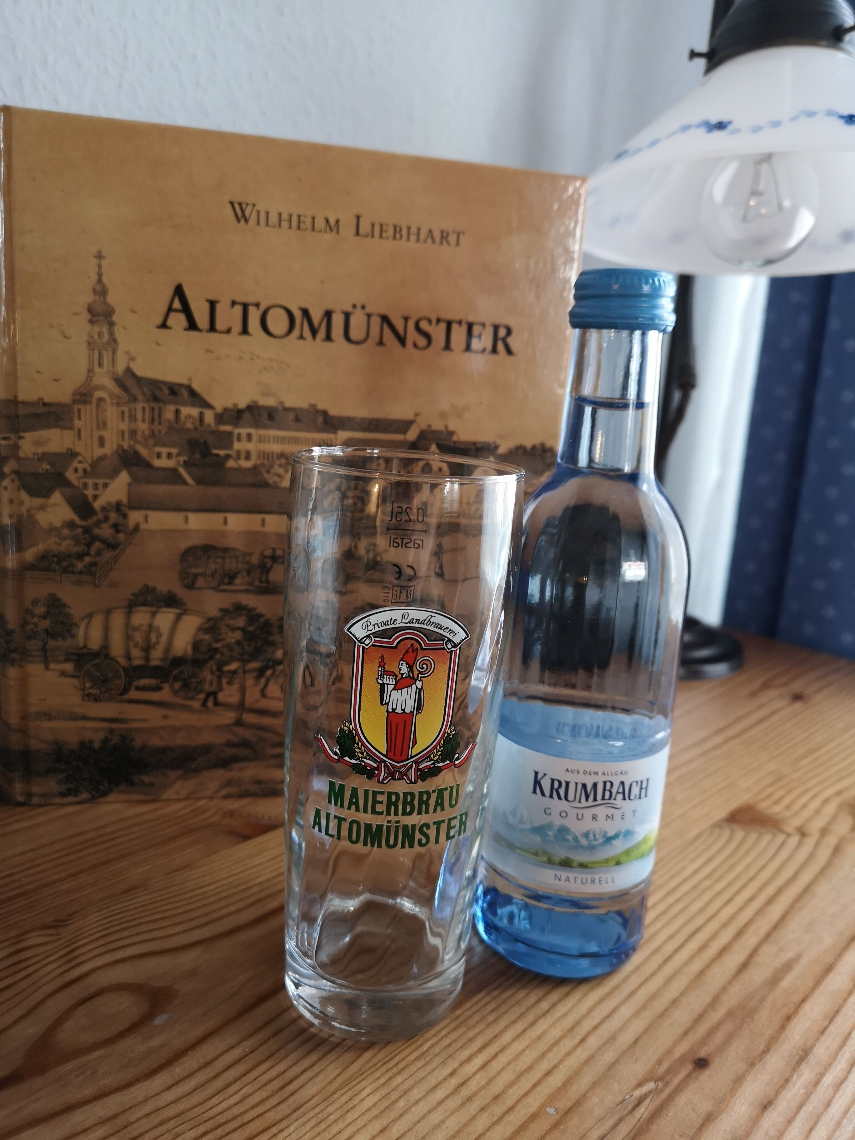 A glass and a bottle of Krumbach liquor on a wooden surface with a box labeled "Wilhelm Liebhart Altomünster" in the background.