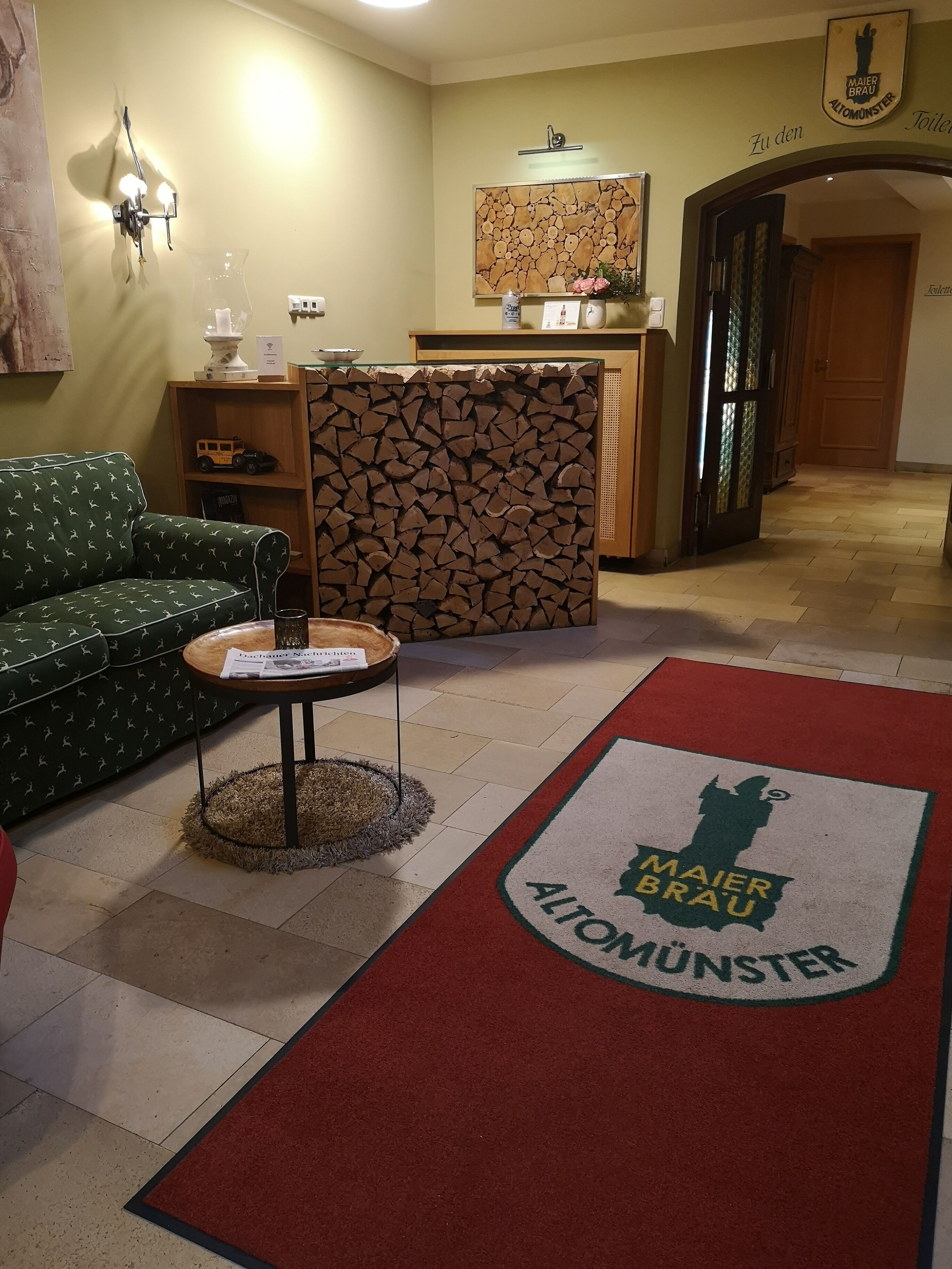 Un hall d’hôtel comprenant un comptoir d’accueil, un canapé à motifs verts, une petite table avec un journal et une moquette rouge ornée du logo de l’hôtel.