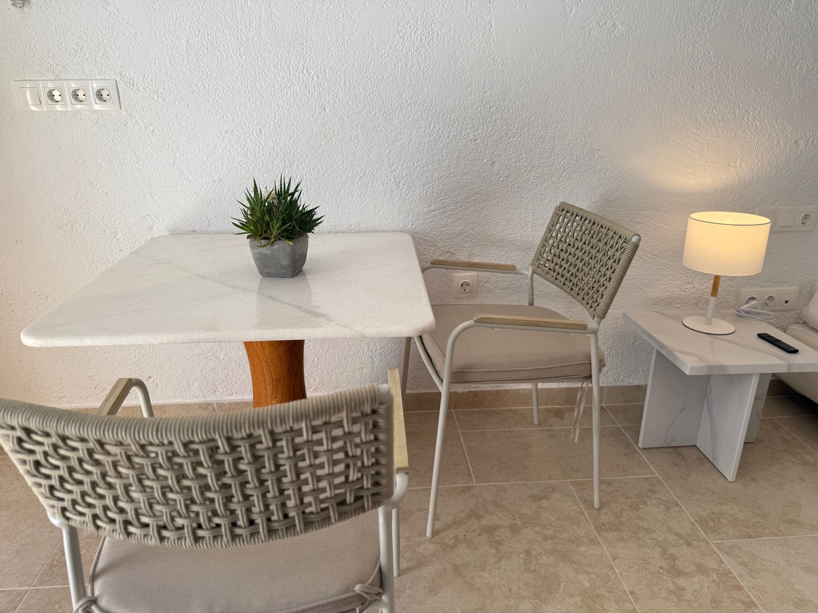 Et lille, godt oplyst rum med et hvidt bord, to stole, en lille potteplante, en lampe og en fjernbetjening på et sengebord.