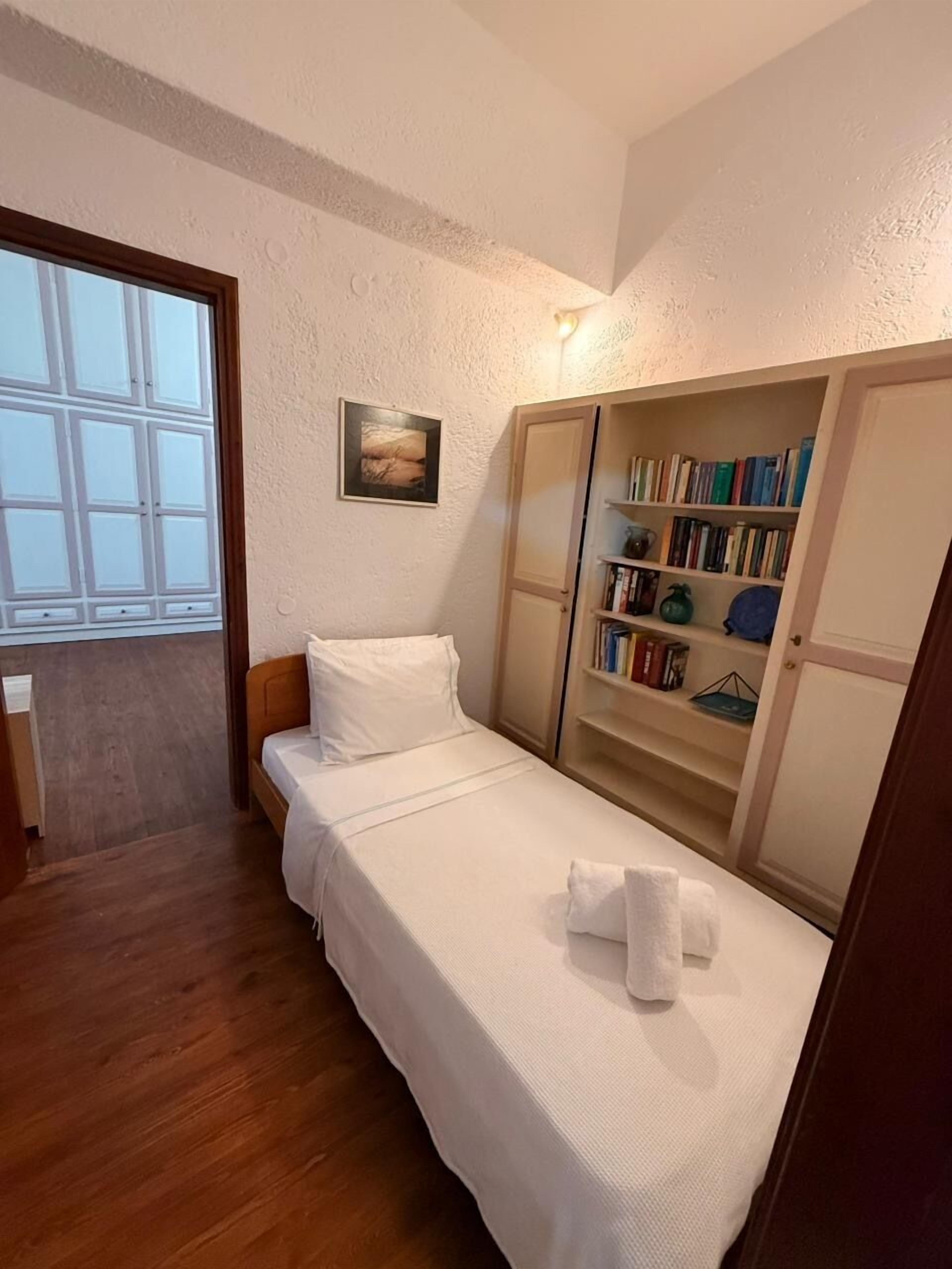 A single bed with a white bedspread, a bedside table, a small bedside lamp, a bookshelf with books, and a window with blinds.