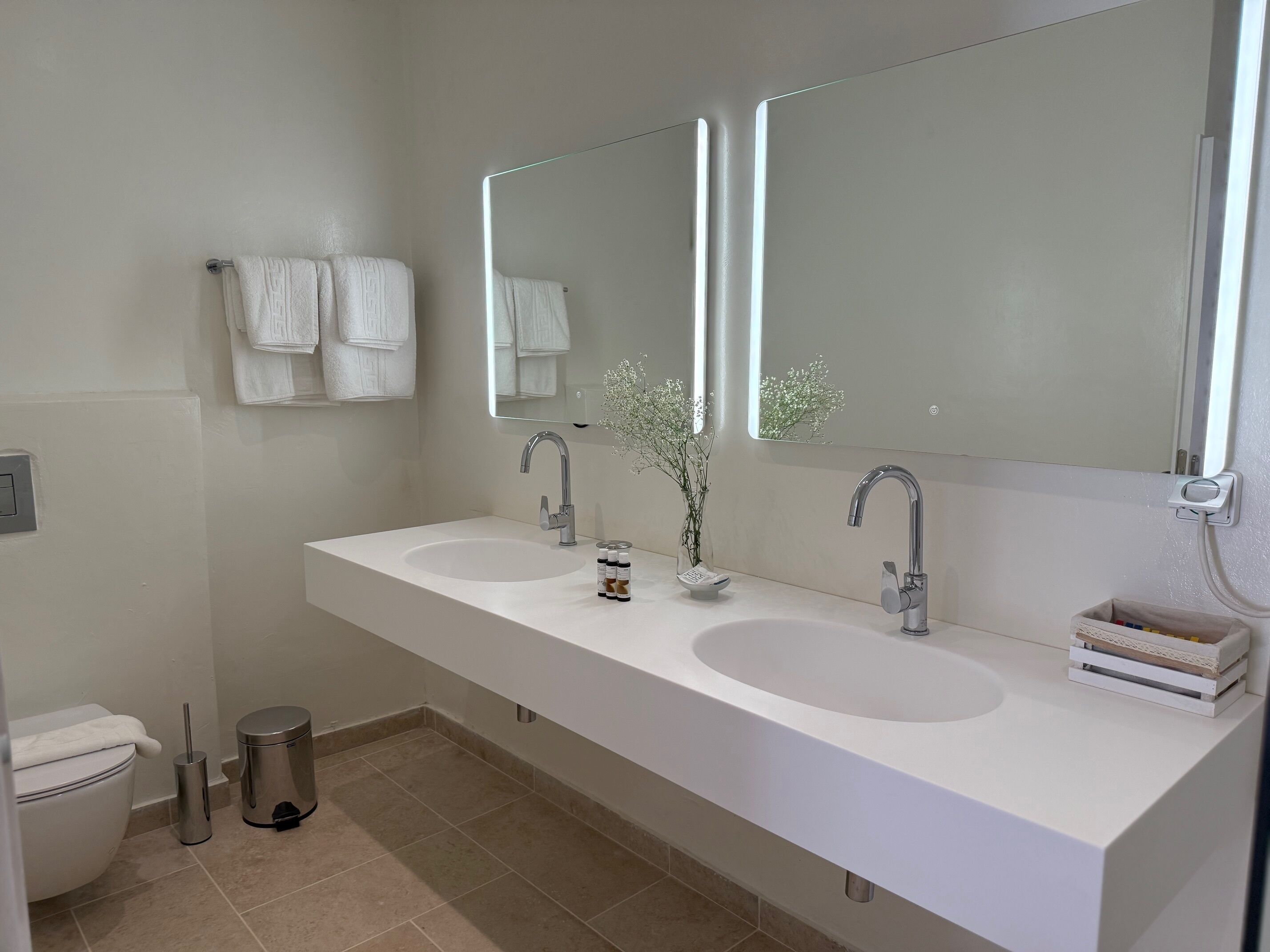 A modern bathroom with two sinks, a toilet, and a trash bin.