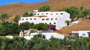A white building with multiple windows and a small bell tower, surrounded by greenery and a hillside.