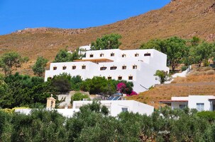 Un edificio blanco con varias ventanas y una pequeña torre de campanas, rodeado de vegetación y una ladera.