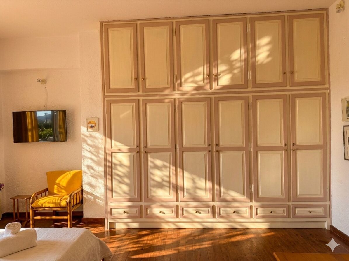 A room with a large wardrobe, a bed with white linens, a yellow armchair, and a painting on the wall.