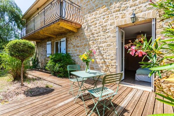 Una terraza de madera con una mesa y sillas, un edificio de piedra con balcón, y una puerta que da a una habitación.