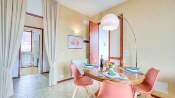 Zona pranzo con un tavolo rotondo, sedie rosa, una lampada e vista su cucina e bagno attraverso una porta aperta.