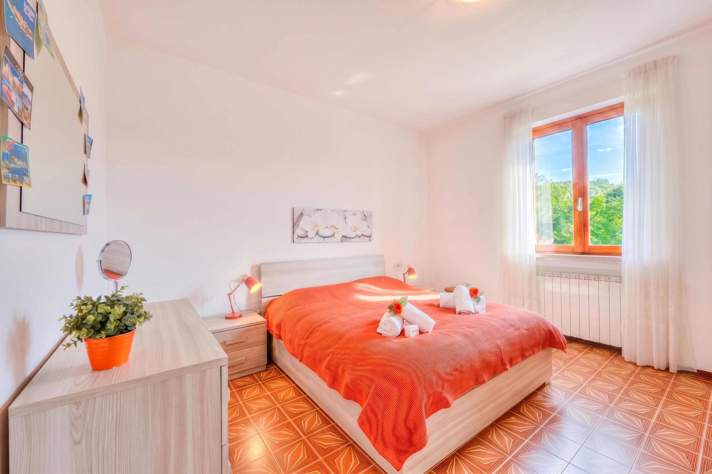 A bedroom with a bed, a nightstand, a potted plant, a window with curtains, and a wall-mounted mirror.