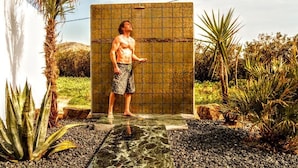 Un homme se tient devant une cabine de douche dont le mur est recouvert de carreaux, entouré de plantes et d’un sol rocheux.