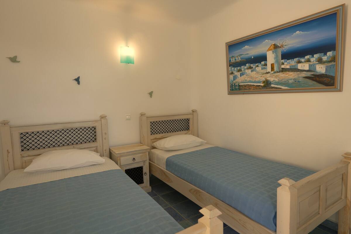 A bedroom with two wooden beds, a nightstand, a wall-mounted lamp, and a painting of a coastal village.