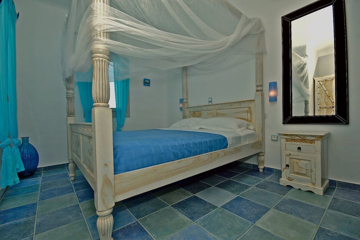 A four-poster bed with a blue bedspread, a nightstand with a mirror, and a blue vase on the floor.