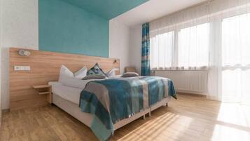 Un dormitorio moderno con cabecera de madera, una cama con manta azul y beige, una mesita de noche y una ventana con cortinas traslúcidas.