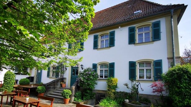 A courtyard with outdoor seating, a building with green shutters, and potted plants. - Landgasthof Büttel (Strullendorf)