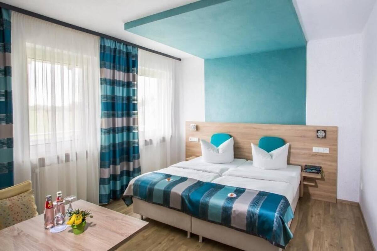 A hotel room with a bed, a bedside table with a lamp, a window with curtains, and a wall with a teal accent.