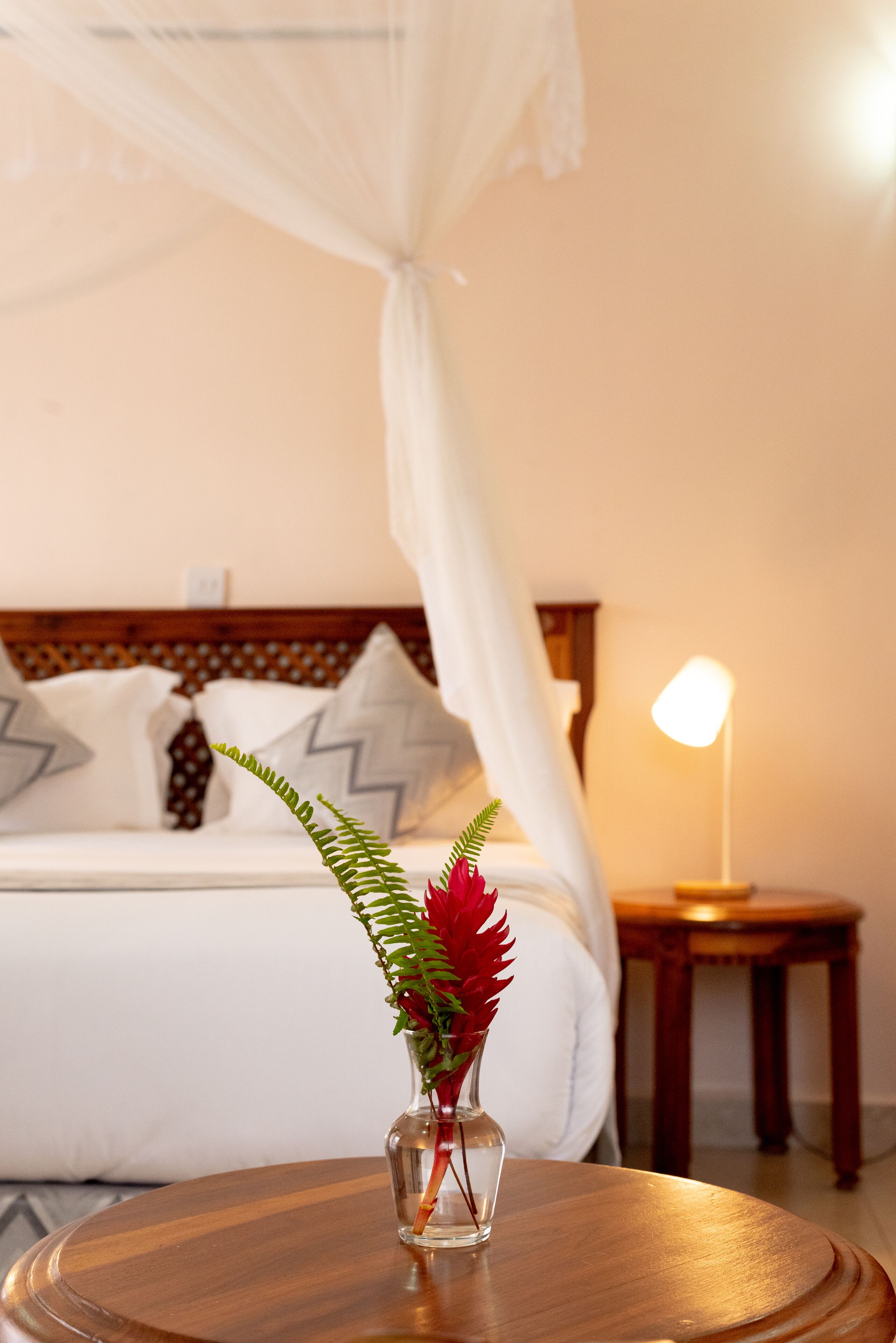 Un lit à baldaquin blanc, une table de chevet avec une lampe et un vase avec une plante, le tout posé sur une petite table ronde en bois.