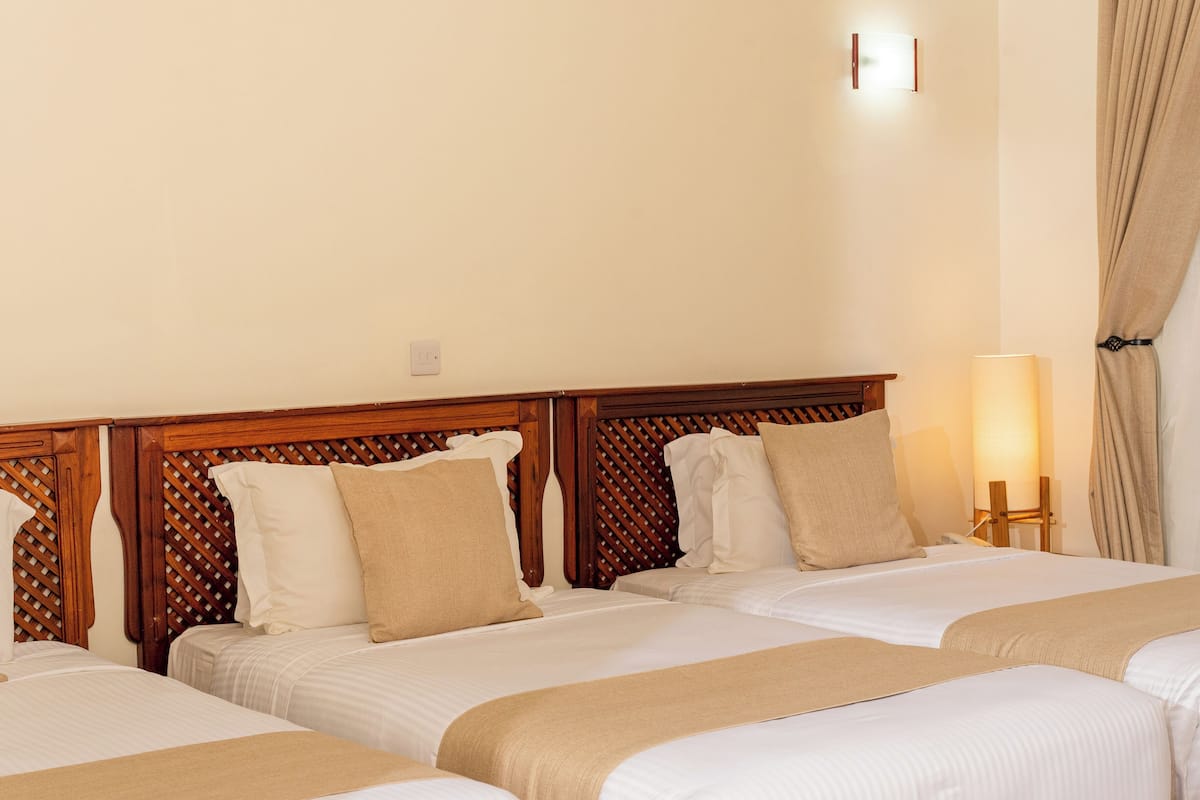 A hotel room with three beds, wooden headboards, beige bedding, and a bedside lamp.